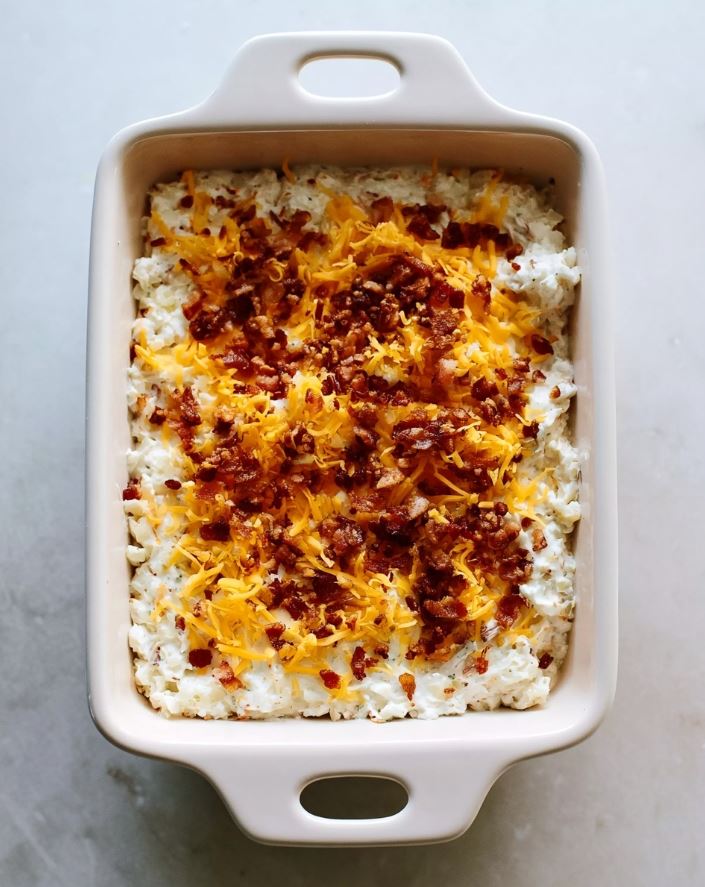Keto Chicken Bacon Ranch Casserole Assembly - casserole filling spread flat in a baking dish, topped with a thick layer of shredded cheddar and crumbled bacon