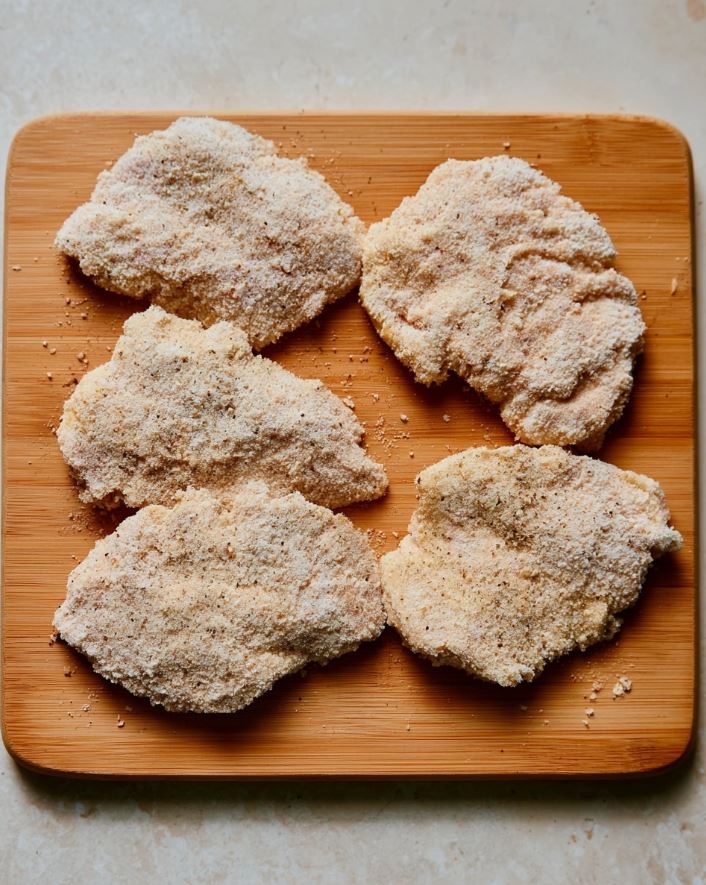 Breading the Chicken - Raw keto chicken parmesan pieces coated in a light, coarse almond flour breading, sitting on a wooden board before frying