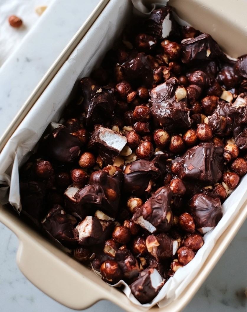 Keto Rocky Road Recipe — dark chocolate mixture pressed into a parchment-lined pan, with a glossy surface highlighting hazelnuts and marshmallows