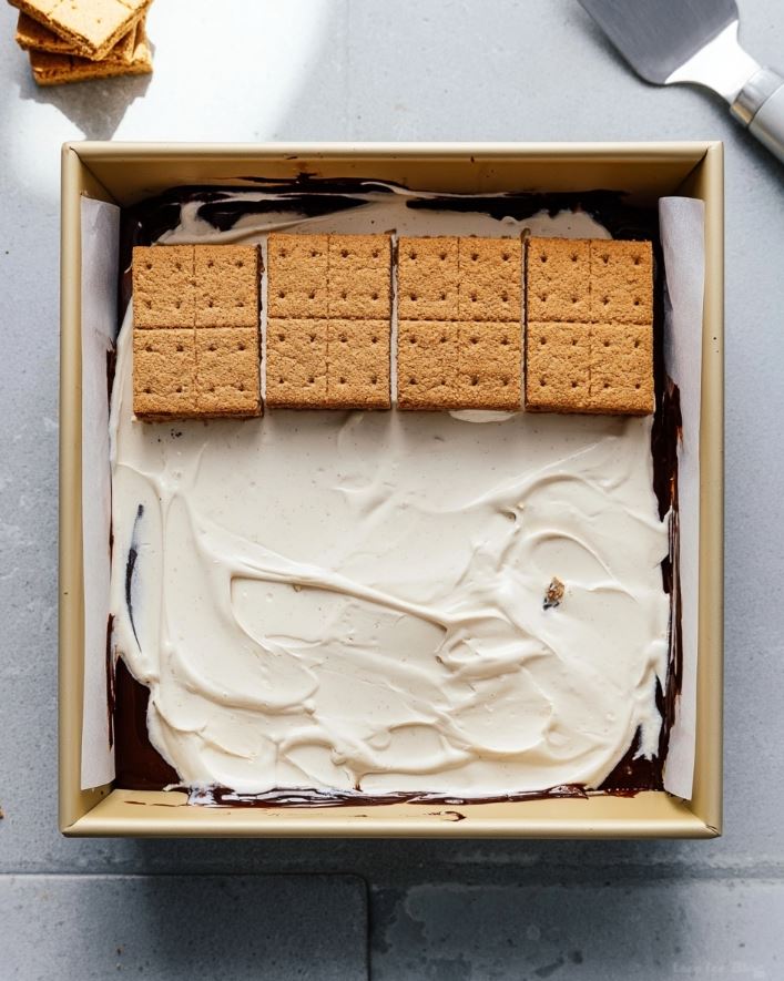 Keto Frozen S'mores Bars - Unfrozen bars in a parchment-lined pan with a smooth cream layer topped by neatly arranged graham crackers.