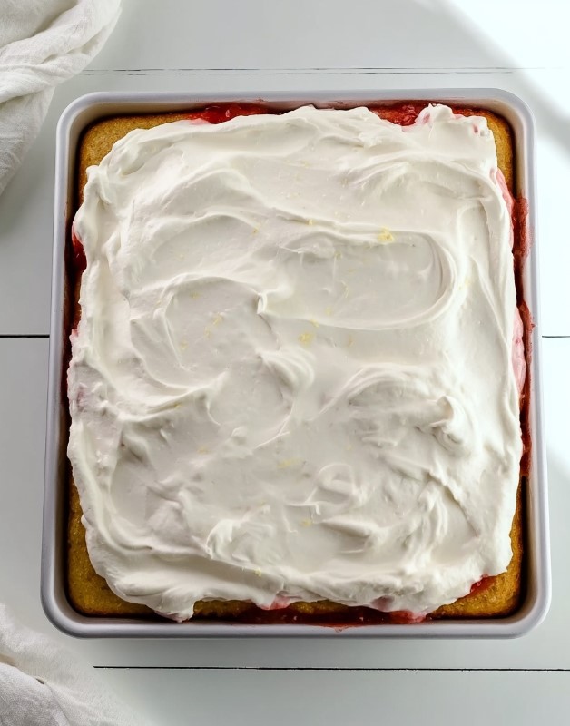 Decorating the cake - A low-carb strawberry shortcake poke cake completely covered with smooth white cream inside a square baking pan