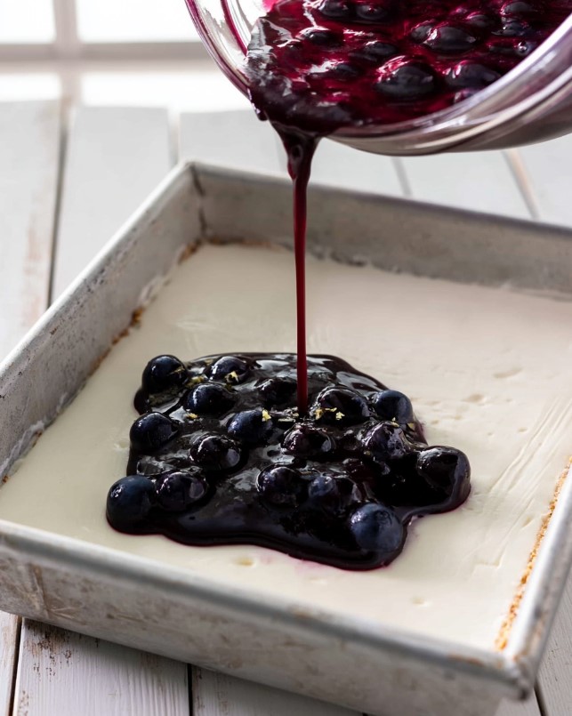 Keto Blueberry Cream Cheese Bars - Pouring glossy blueberry sauce over the cream layer in the keto blueberry pecan dessert