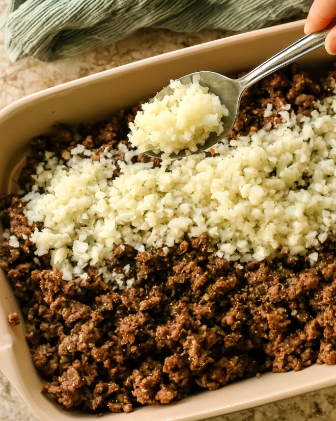 Keto Hamburger Casserole — Spreading cauliflower rice over seasoned ground beef in a ceramic baking dish while assembling the layers.