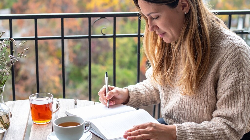 Woman writing in a notebook beside cups of tea while sitting at an outdoor table.