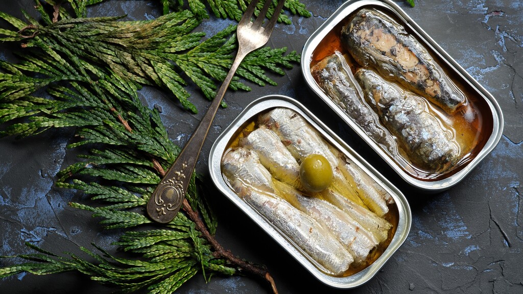 Open tins of sardines in olive oil on a dark surface with herbs and a fork.