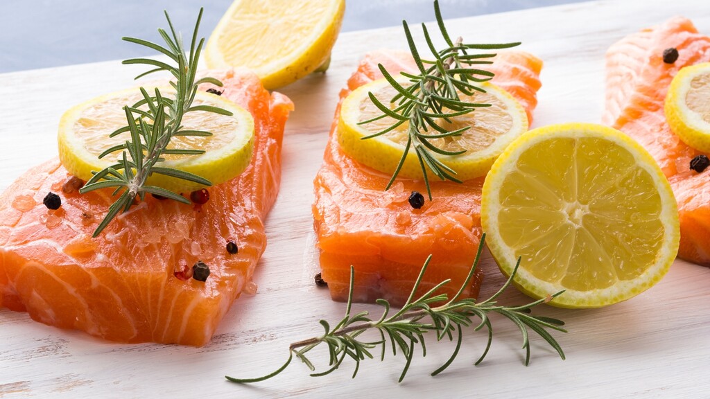 Raw salmon fillets with lemon slices, rosemary sprigs, and black pepper on a white cutting board.