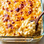 Wooden spoon lifting a creamy portion of baked macaroni and cheese from a glass baking dish with a golden brown top