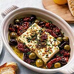 Warm baked Mediterranean feta dip in a ceramic baking dish surrounded by mixed olives and sun-dried tomatoes with toasted baguette slices