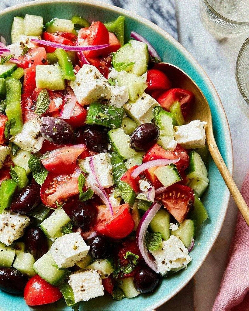 Finished Greek salad ready to serve in a green bowl, topped with large chunks of feta, olives, and fresh mint