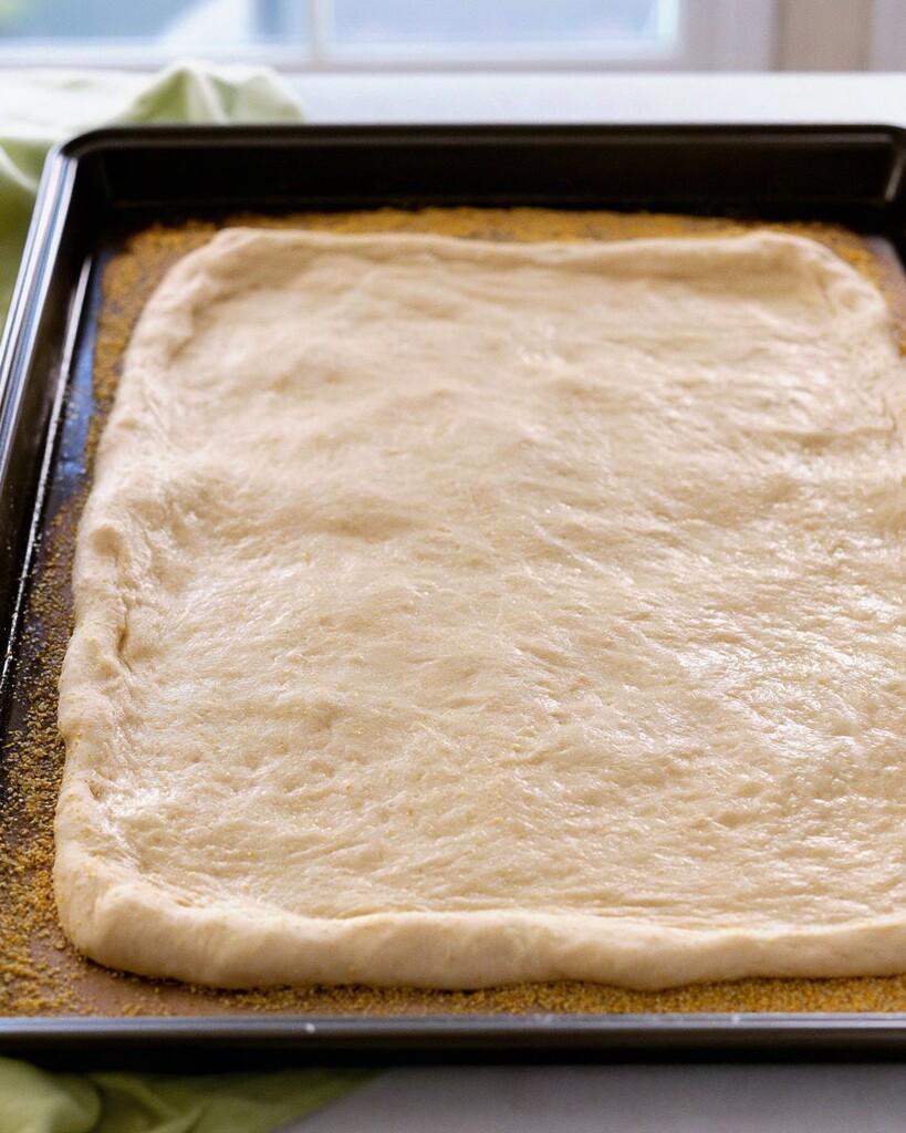 Unbaked pizza dough stretched into a rectangle in a black pan dusted with cornmeal