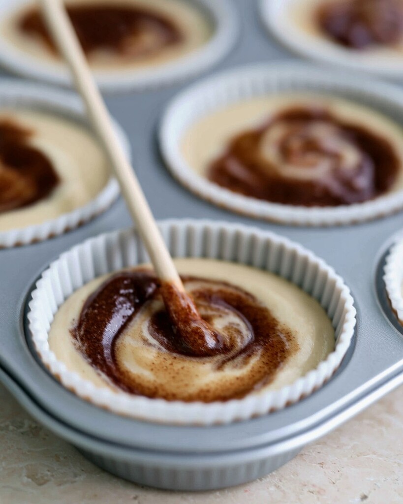 A wooden skewer marbling brown cinnamon paste into muffin batter inside white liners