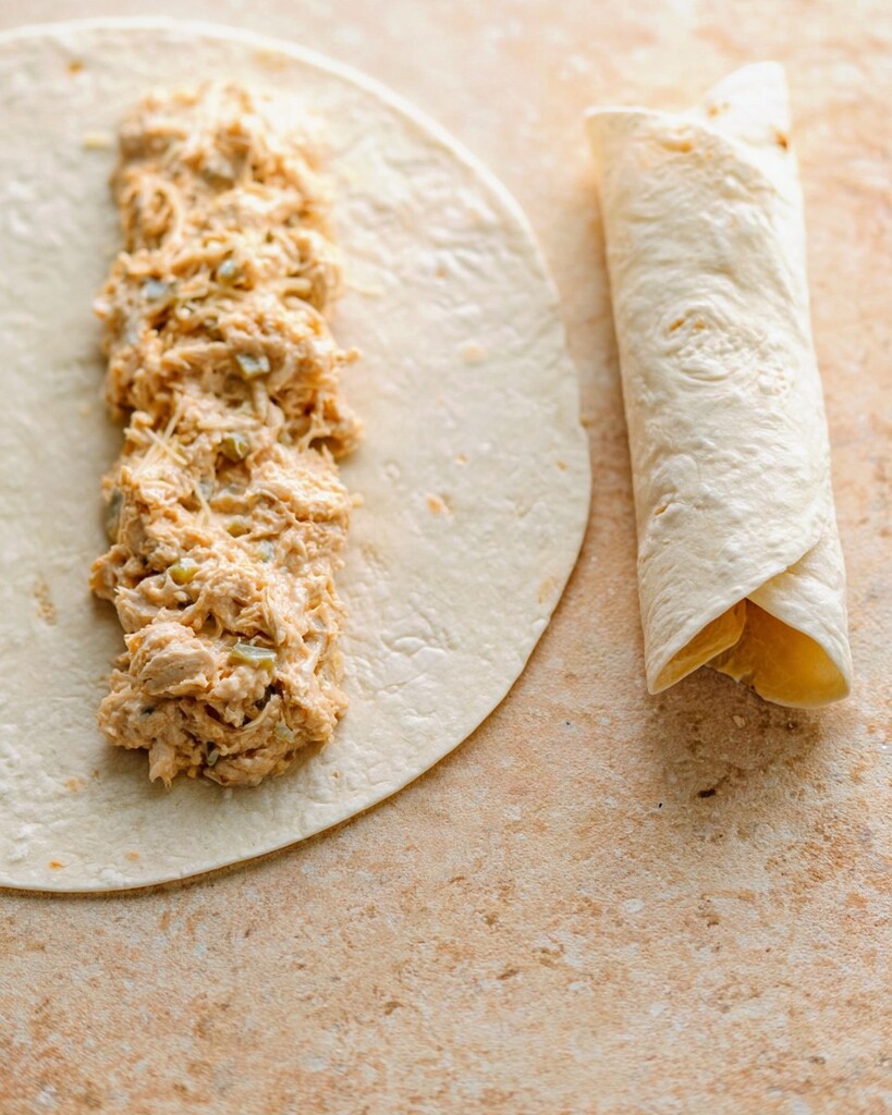 Creamy chicken enchilada filling distributed in a line down the center of a flour tortilla next to a tightly rolled enchilada