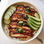 Sliced crispy chicken katsu arranged in a bowl with rice, avocado, cucumbers, and sesame seeds