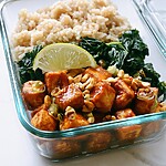 Glass container holding glazed tofu cubes, brown rice, chopped kale, peanuts, and a lime wedge