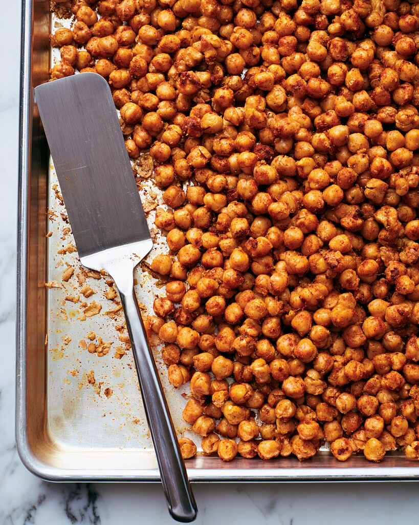 Golden brown roasted chickpeas on a metal pan being tossed with paprika and garlic seasoning using a spatula