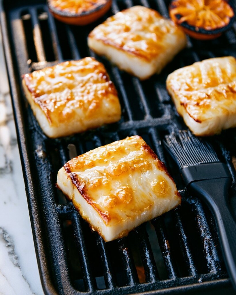 White fish fillets cooking skin-side down on a black grill grate with a basting brush and browned orange halves nearby