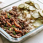 Glass meal prep bowl filled with ground beef, rice, pickled cucumbers, scallions, and a creamy orange sauce