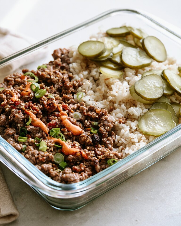 Glass meal prep bowl filled with ground beef, rice, pickled cucumbers, scallions, and a creamy orange sauce