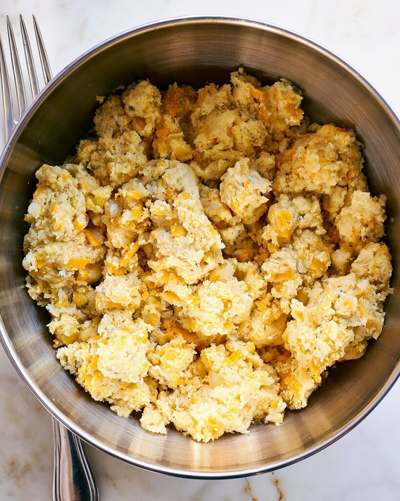 Crumbly biscuit dough mixed with shredded cheddar cheese inside a metal mixing bowl