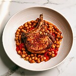 Seared bone-in pork chop resting on a bed of roasted chickpeas and red peppers in a white plate