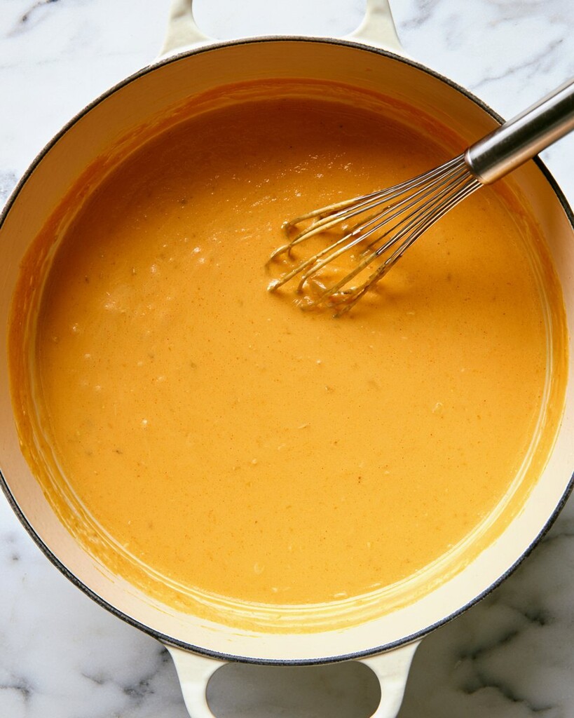 Whisking a vibrant orange cheese sauce in a heavy pot until smooth and velvety