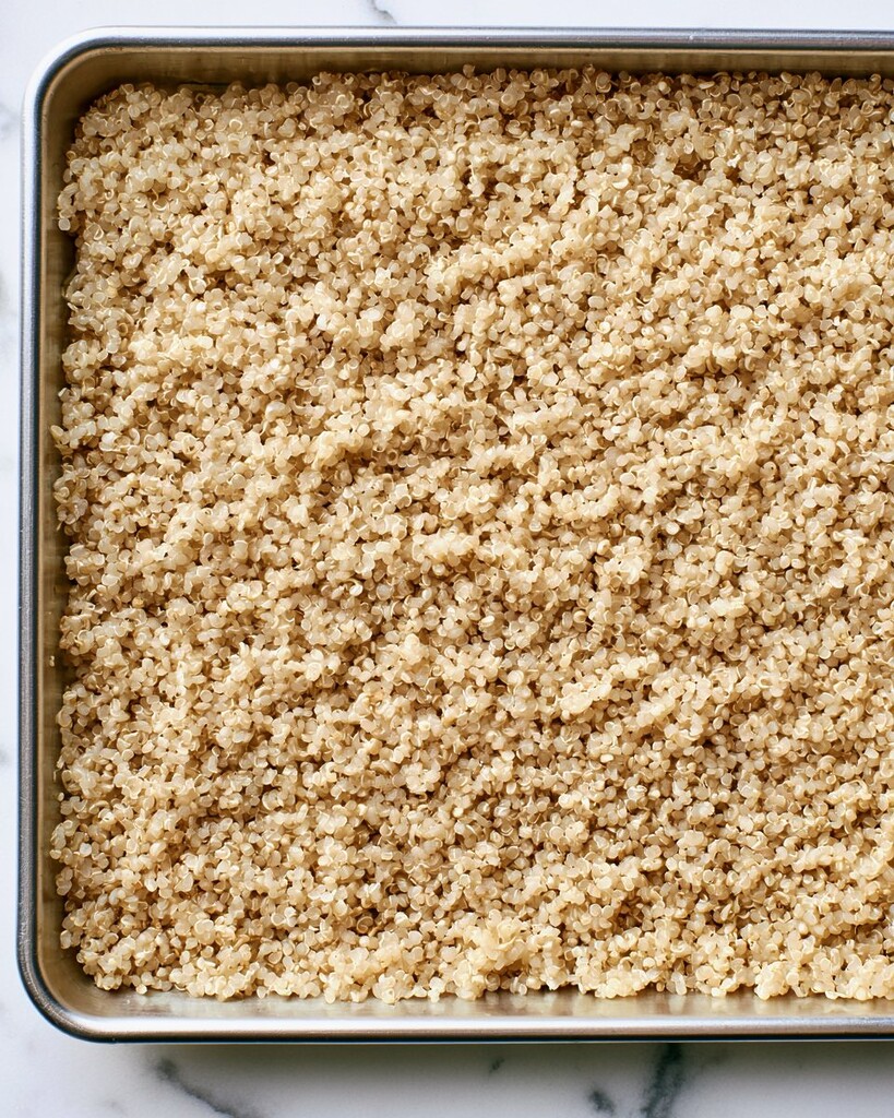 Cooked white quinoa spread across a wide metal baking sheet to cool with distinct fluffy grains