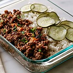 A glass serving bowl displaying the finished Korean beef over rice with bright orange sauce and pickles