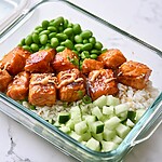An open meal prep container with rice, salmon, edamame, and cucumber, topped with a drizzle of spicy mayo and sesame seeds