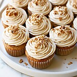 A fully decorated banana cupcake on a white plate showing a light spiral of frosting and chopped nuts