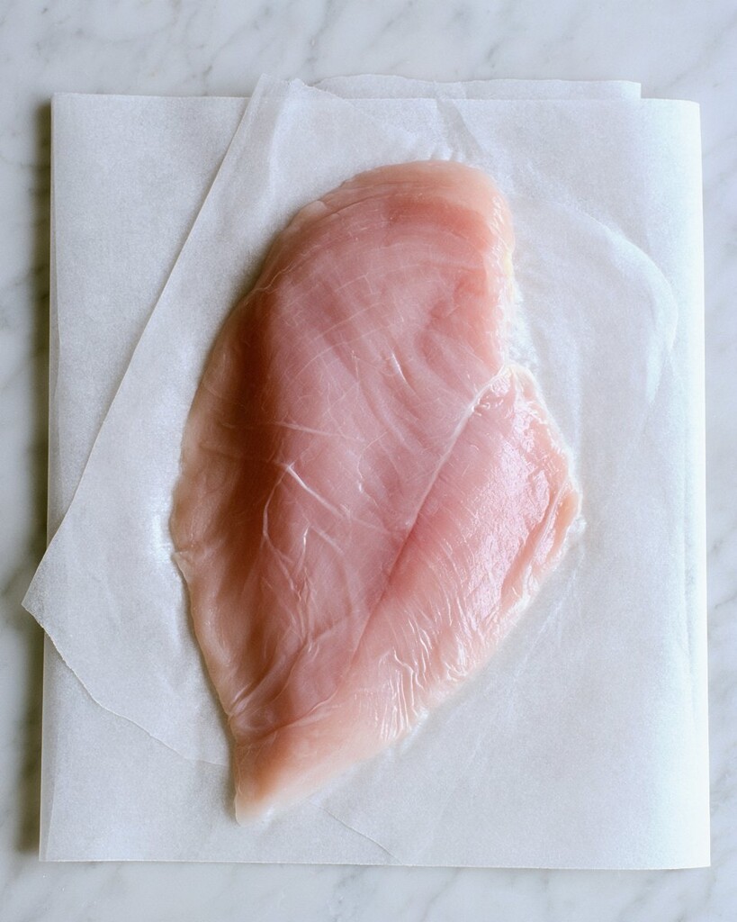 Raw chicken breast pounded flat on white parchment paper