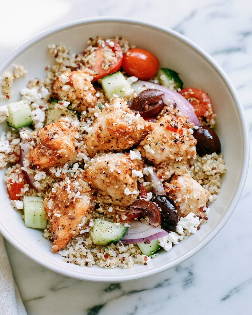 Assembling a bowl with seared chicken, diced vegetables, olives, and feta over quinoa