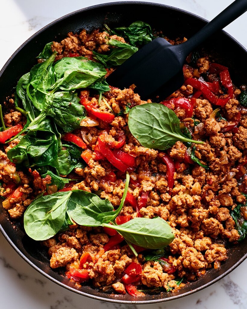 Fresh spinach leaves added to glazed ground turkey and red bell peppers in a skillet