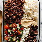 Glass meal prep container being filled with crispy za'atar beef, hummus, rice, and fresh salad