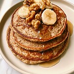 Stack of browned whole wheat pancakes topped with sliced bananas and chopped walnuts, surrounded by maple syrup