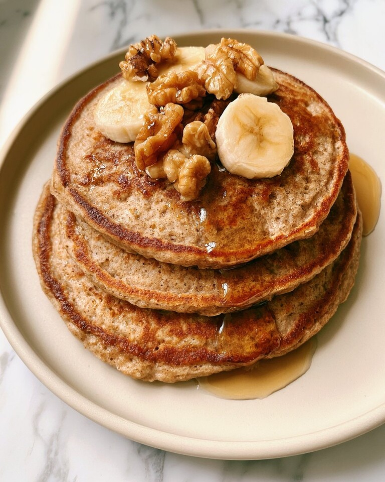 Stack of browned whole wheat pancakes topped with sliced bananas and chopped walnuts, surrounded by maple syrup
