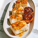 Golden glazed halibut fillets on an oval plate garnished with sliced basil and charred orange halves
