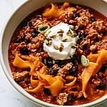 A serving of turkey chili with butternut squash noodles in a round bowl, garnished with sour cream, pumpkin seeds, and sliced scallions.