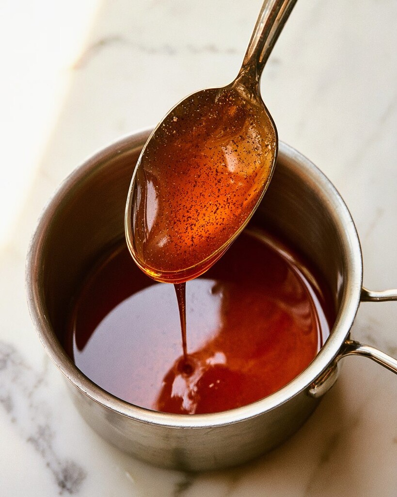 Spoon lifted from a small saucepan showing a glossy thick hot honey pepper sauce clinging to the metal