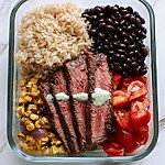 A glass meal prep bowl containing brown rice, black beans, charred corn, red onions, tomatoes, and sliced steak topped with green cilantro yogurt crema