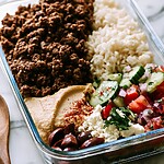 1 glass meal prep containers filled with crispy beef, hummus, rice, feta, and cucumber salad