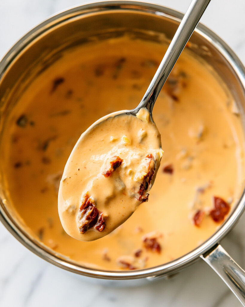 Thick pale orange cream sauce with chopped sun-dried tomatoes simmering in a metal saucepan