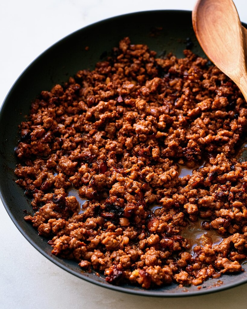 Ground beef cooking in a dark skillet with a wooden spoon, coated in a dark glossy glaze