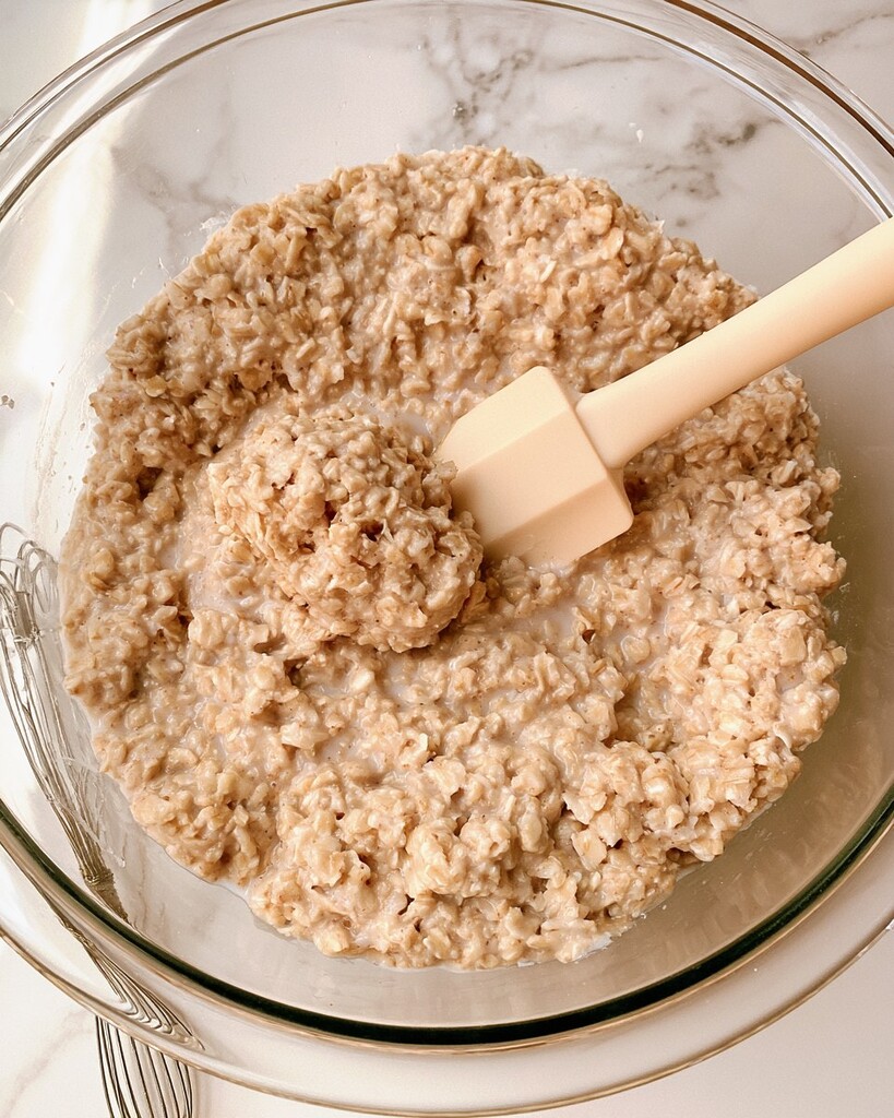 Wet soaked rolled oats mixed with almond milk in a glass bowl with a silicone spatula