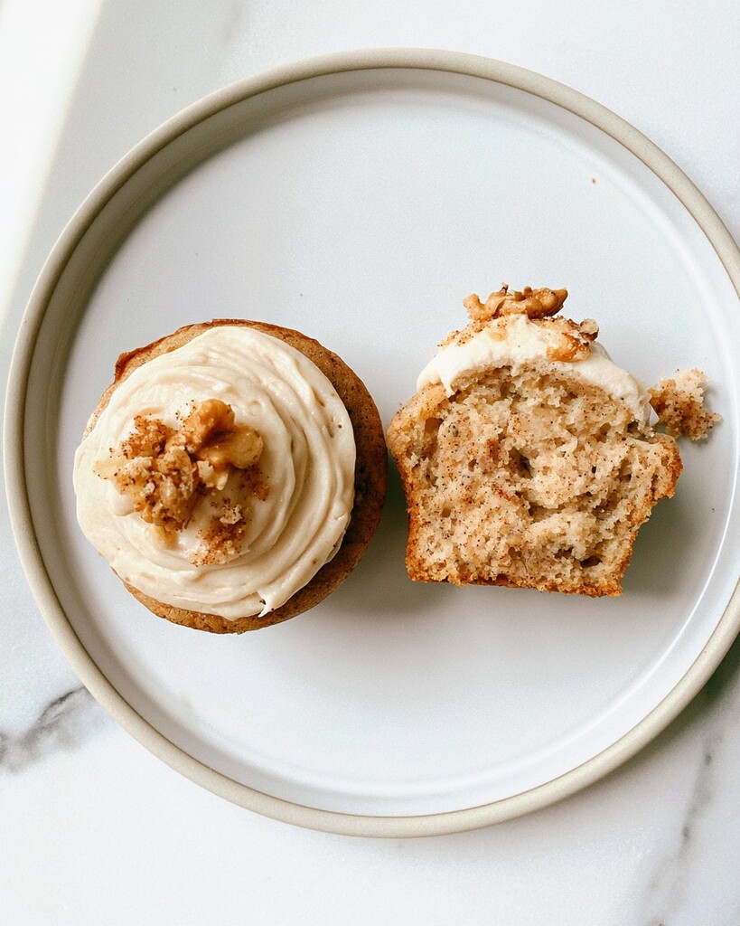 Two frosted banana cupcakes on a white plate with one sliced in half to show the internal crumb
