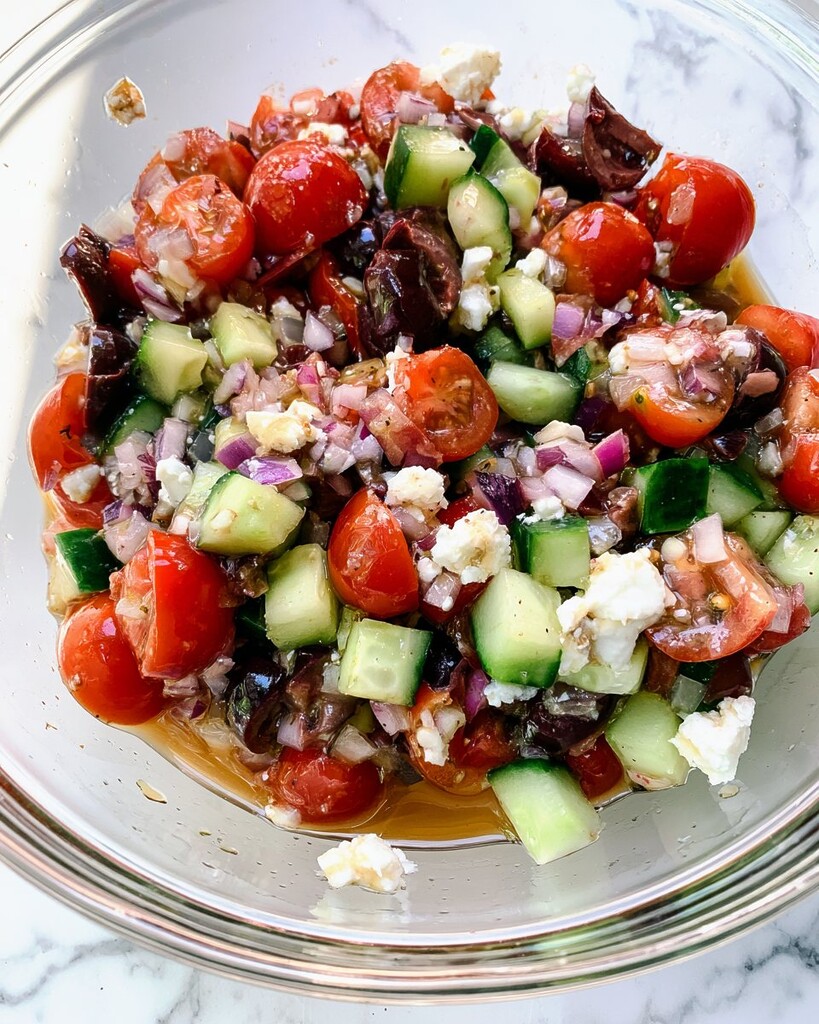 Diced cucumbers, cherry tomatoes, red onions, olives, and feta mixed in a glass bowl