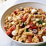 A wide serving bowl filled with a glistening Italian pasta salad featuring rotini, chicken, salami, mozzarella pearls, and chopped vegetables