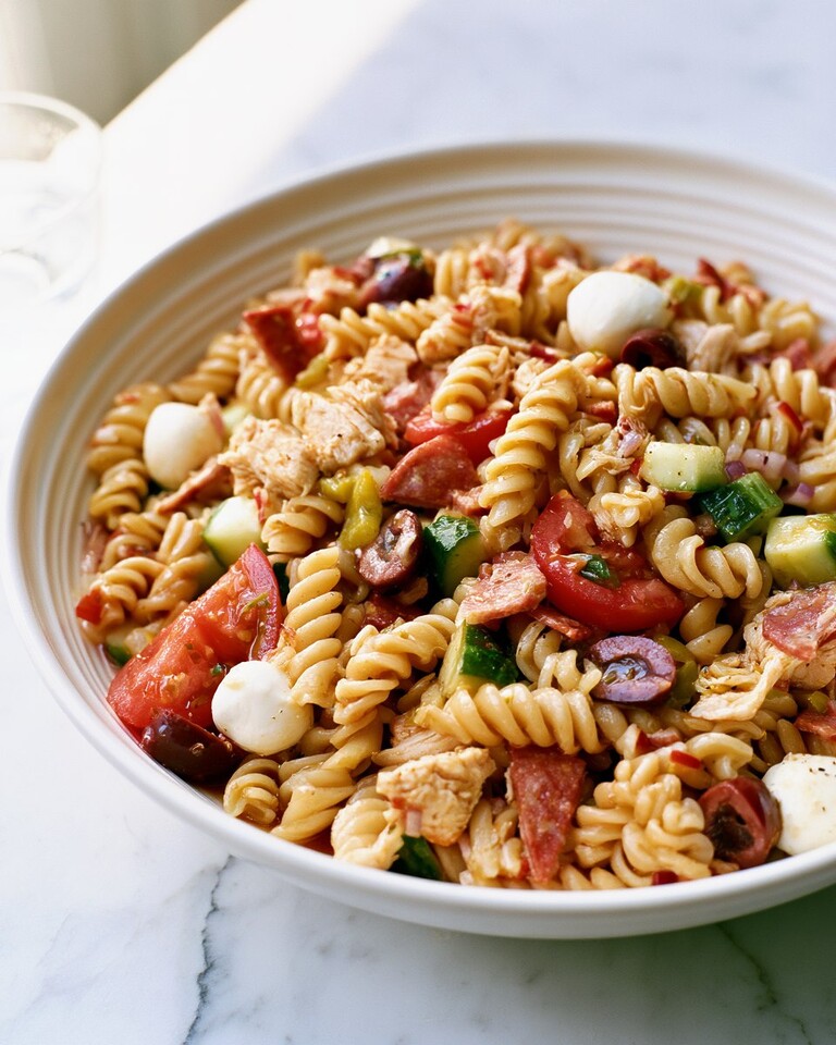 A wide serving bowl filled with a glistening Italian pasta salad featuring rotini, chicken, salami, mozzarella pearls, and chopped vegetables