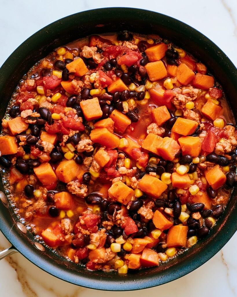 Turkey, sweet potatoes, beans, and corn simmering in a red tomato sauce in a skillet