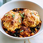 A white bowl filled with roasted cauliflower and black bean chili, topped with two golden cheddar biscuits and sliced green scallions