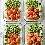 Four glass meal prep containers lined up, filled with white rice, glazed salmon cubes, diced cucumbers, and edamame